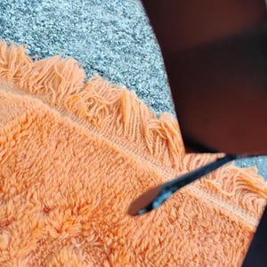 May include: Close-up of an orange fringed rug with a textured surface. The rug is a warm, inviting color and has a soft, plush appearance. The rug is partially visible, with a dark object in the upper right corner.