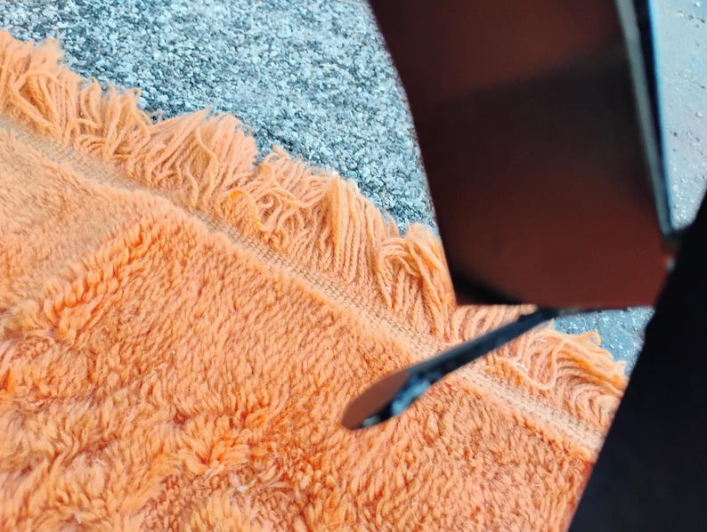 May include: Close-up of an orange fringed rug with a textured surface. The rug is a warm, inviting color and has a soft, plush appearance. The rug is partially visible, with a dark object in the upper right corner.