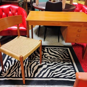 May include: A mid-century modern desk with three drawers, a woven chair, and a zebra-print rug. The desk and chair are made of wood. Two red chairs are in the background. The rug is black and white.
