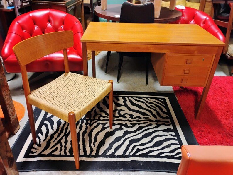 May include: A mid-century modern desk with three drawers, a woven chair, and a zebra-print rug. The desk and chair are made of wood. Two red chairs are in the background. The rug is black and white.