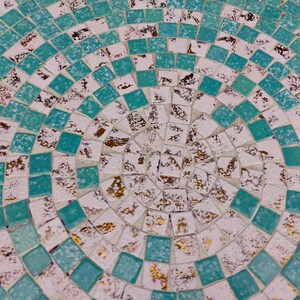 Op de afbeelding: Een cirkelvormig moza&iuml;ekpatroon met een witte achtergrond en turquoise blauwe tegels. Het midden van het patroon is een cirkel van gouden en witte tegels.