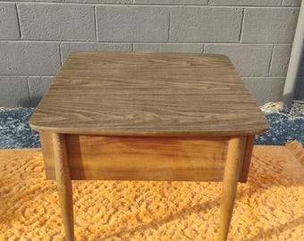 May include: A mid-century modern side table with a rectangular top and tapered legs. The table has a wood grain finish and a drawer. The legs are approximately 18 inches tall. The table is set on an orange shag carpet.