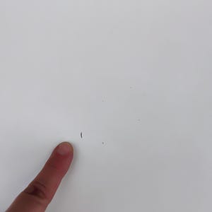 May include: A close-up of a white surface with a small dark mark and a finger pointing towards it. The surface is smooth and clean, with a few tiny imperfections.