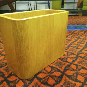 Puede incluir: Un cesto de basura rectangular de madera con acabado de roble claro. El cesto tiene esquinas redondeadas y una superficie lisa. El interior está abierto y vacío. El cesto está sobre una alfombra estampada.