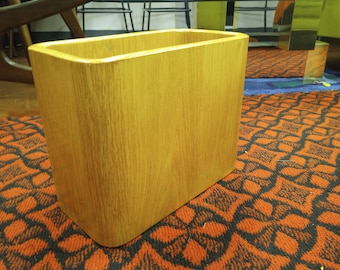 Könnte beinhalten: Ein rechteckiger Holz-Abfalleimer mit hellem Eichenholz-Finish. Der Abfalleimer hat abgerundete Ecken und eine glatte Oberfläche. Der Innenraum ist offen und leer. Der Abfalleimer steht auf einem gemusterten Teppich.