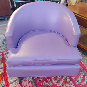 May include: A purple upholstered armchair with a rounded back and a tufted seat cushion. The chair has a simple design and is sitting on a colorful rug.