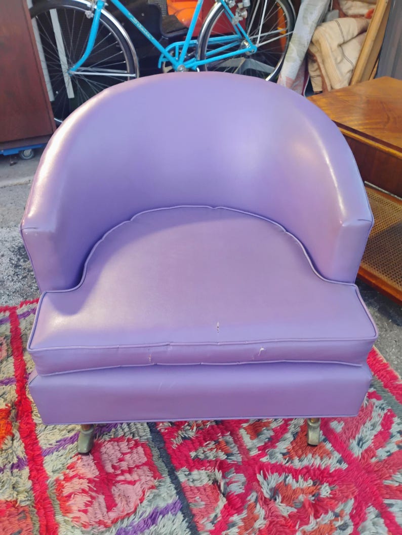 May include: A purple upholstered armchair with a rounded back and a tufted seat cushion. The chair has a simple design and is sitting on a colorful rug.