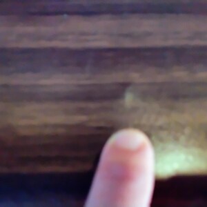 May include: Close-up of a finger pointing towards a dark brown wooden surface. The wood grain is visible, with variations in color and texture. The lighting is soft, highlighting the natural patterns.