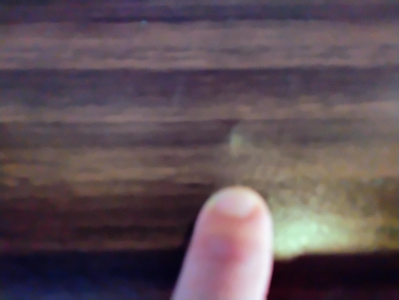 May include: Close-up of a finger pointing towards a dark brown wooden surface. The wood grain is visible, with variations in color and texture. The lighting is soft, highlighting the natural patterns.