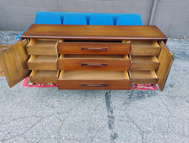 May include: A long wooden dresser with a smooth top surface, featuring three central drawers with horizontal handles and two side cabinets with three internal drawers each. The cabinet doors are open, revealing the interior storage compartments.