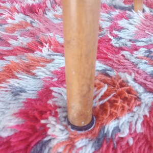 May include: Close-up of a wooden chair leg with a black ring at the bottom. The chair leg is standing on a colorful rug with red, white, and gray patterns.