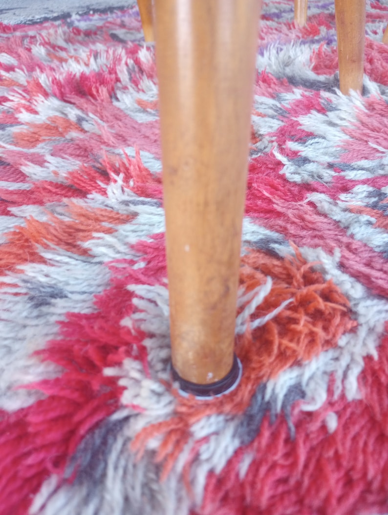 May include: Close-up of a wooden chair leg with a black ring at the bottom. The chair leg is standing on a colorful rug with red, white, and gray patterns.