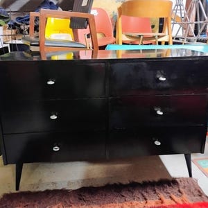 May include: A black six-drawer dresser with silver knobs and tapered legs. The dresser has a glossy finish and is set against a backdrop of various furniture pieces, suggesting a vintage or antique aesthetic.