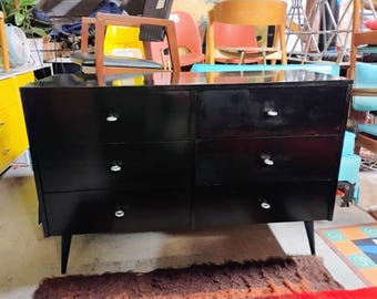 Könnte beinhalten: Eine schwarze Kommode mit sechs Schubladen, silbernen Knöpfen und konischen Beinen. Die Kommode hat eine glänzende Oberfläche. Sie steht vor einer Kulisse aus verschiedenen Möbelstücken, was auf eine Vintage- oder antike Ästhetik hindeutet.