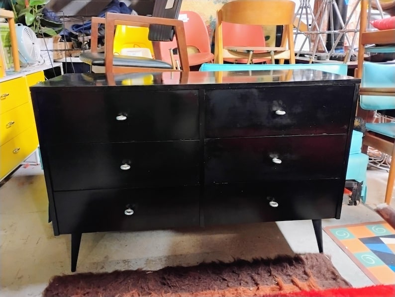 May include: A black six-drawer dresser with silver knobs and tapered legs. The dresser has a glossy finish and is set against a backdrop of various furniture pieces, suggesting a vintage or antique aesthetic.