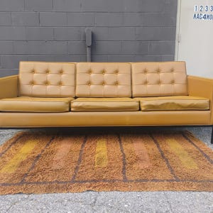 May include: A three-seat sofa upholstered in mustard yellow leather featuring a tufted backrest with buttons, three rectangular seat cushions, and a black metal frame with four straight legs, positioned above a textured brown and orange rug.
