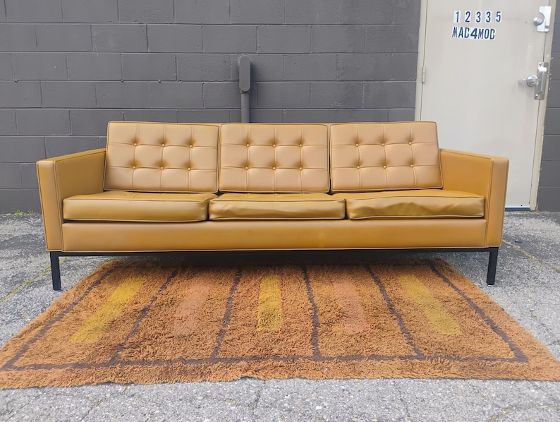 May include: A three-seat sofa upholstered in mustard yellow leather featuring a tufted backrest with buttons, three rectangular seat cushions, and a black metal frame with four straight legs, positioned above a textured brown and orange rug.