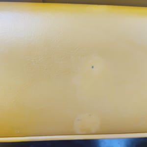 May include: A smooth, mustard yellow vinyl surface with a subtle leather-like texture. The material features a small, dark blue circular mark near the center and a faint, lighter-colored circular discoloration towards the bottom edge of the frame.
