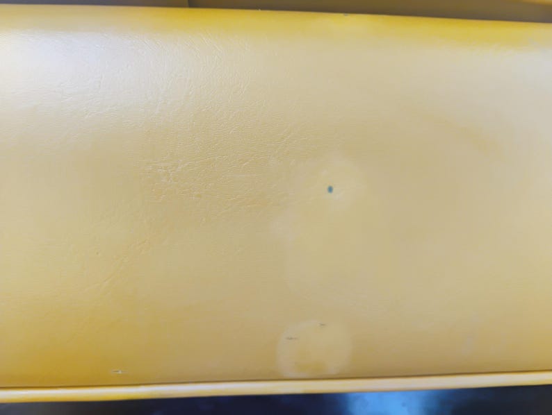May include: A smooth, mustard yellow vinyl surface with a subtle leather-like texture. The material features a small, dark blue circular mark near the center and a faint, lighter-colored circular discoloration towards the bottom edge of the frame.