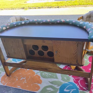 May include: A wooden poker table with a dark tabletop and a green padded rail with colorful designs. The table has a cabinet with storage and a floral patterned rug underneath. Two chairs are visible in the background.