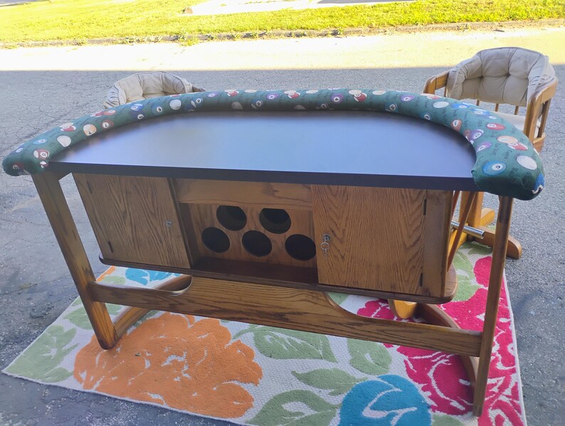 May include: A wooden poker table with a dark tabletop and a green padded rail with colorful designs. The table has a cabinet with storage and a floral patterned rug underneath. Two chairs are visible in the background.