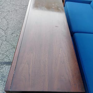 May include: A long, rectangular wooden sideboard with a dark brown finish and visible wood grain patterns. The top surface is flat and smooth, showing a polished texture with some light surface wear and subtle variations in the wood tone.