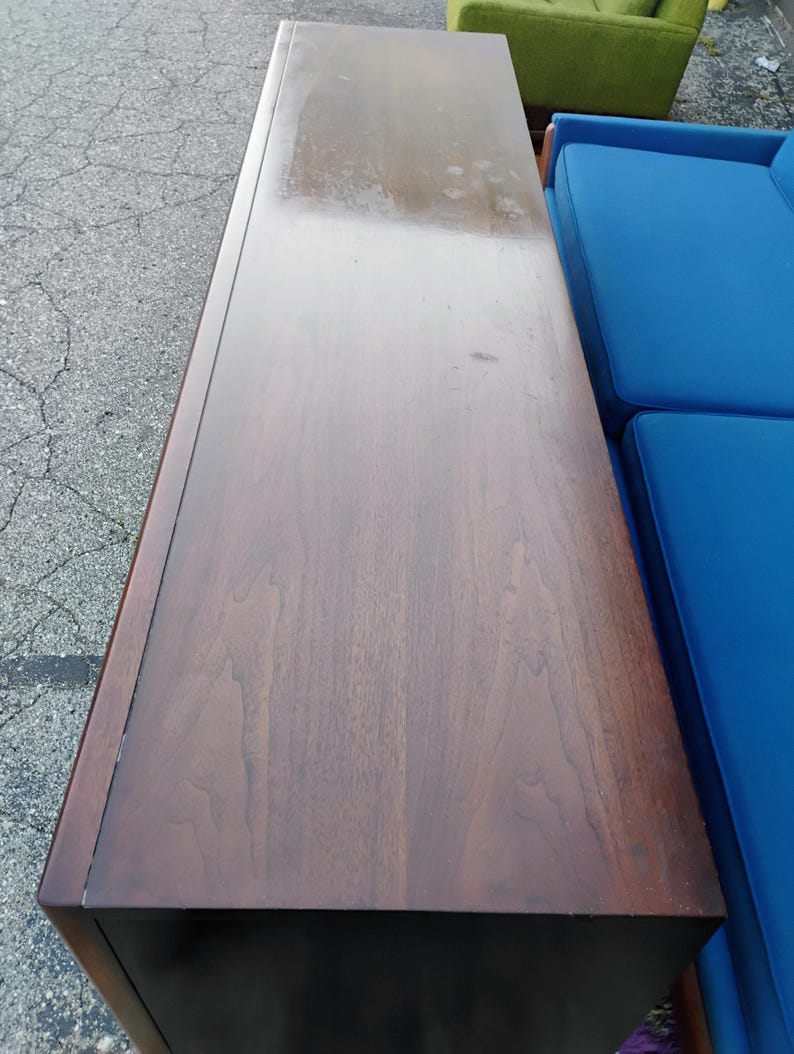 May include: A long, rectangular wooden sideboard with a dark brown finish and visible wood grain patterns. The top surface is flat and smooth, showing a polished texture with some light surface wear and subtle variations in the wood tone.