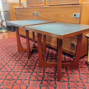 May include: Two square side tables featuring dark gray tops and brown wooden frames with curved, U-shaped legs. The tables have a clean, geometric design with smooth surfaces and sturdy bases, positioned side by side on a patterned red and black rug.