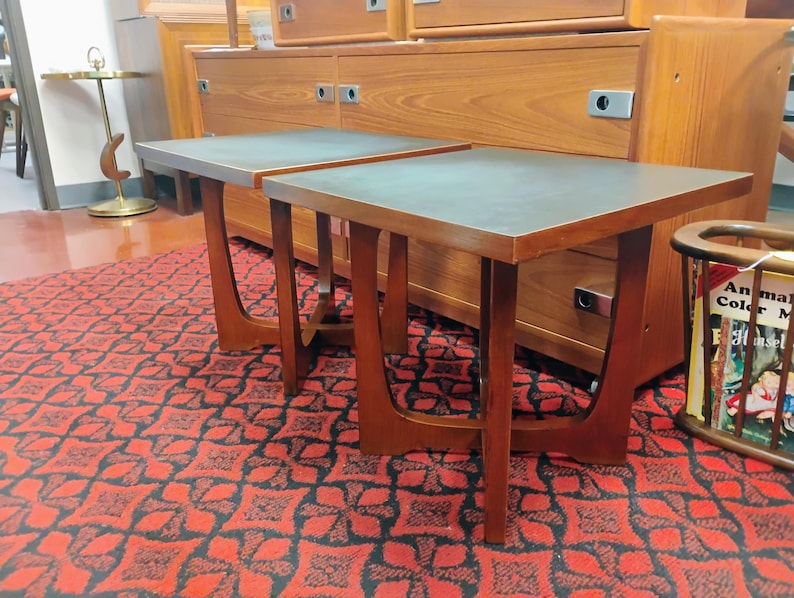 May include: Two square side tables featuring dark gray tops and brown wooden frames with curved, U-shaped legs. The tables have a clean, geometric design with smooth surfaces and sturdy bases, positioned side by side on a patterned red and black rug.