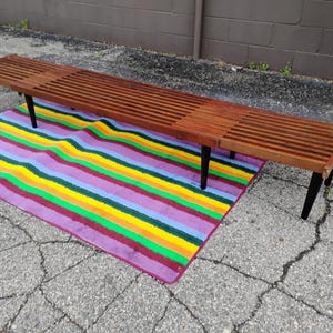 K&ouml;nnte beinhalten: Eine lange Holzbank mit Lamellendesign und schwarzen Beinen. Die Bank ist etwa 183 cm lang und steht auf einem bunten gestreiften Teppich mit lila, gelben, gr&uuml;nen und blauen Streifen.