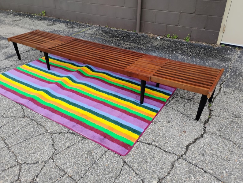 K&ouml;nnte beinhalten: Eine lange Holzbank mit Lamellendesign und schwarzen Beinen. Die Bank ist etwa 183 cm lang und steht auf einem bunten gestreiften Teppich mit lila, gelben, gr&uuml;nen und blauen Streifen.