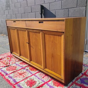 Op de afbeelding: Een rechthoekig houten dressoir met een warme bruine afwerking, twee horizontale lades aan de bovenzijde en vier kastdeuren eronder. Het meubel heeft een vlak bovenblad en een strakke, lineaire constructie met verzonken handgrepen.