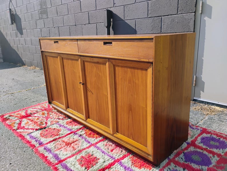 Op de afbeelding: Een rechthoekig houten dressoir met een warme bruine afwerking, twee horizontale lades aan de bovenzijde en vier kastdeuren eronder. Het meubel heeft een vlak bovenblad en een strakke, lineaire constructie met verzonken handgrepen.