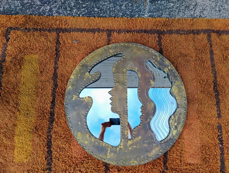 K&ouml;nnte beinhalten: Ein runder, messingfarbener Spiegel mit einem einzigartigen Design, das zwei Profilsilhouetten und ein wellenf&ouml;rmiges Muster zeigt. Der Spiegel reflektiert einen blauen Himmel und liegt auf einem strukturierten, orangefarbenen Teppich.