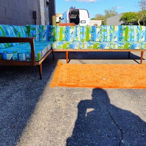 May include: A mid-century modern sectional sofa with a wooden frame and patterned cushions in shades of blue, green, and white. An orange shag rug is in front of the sofa. The sofa is outdoors on a sunny day.