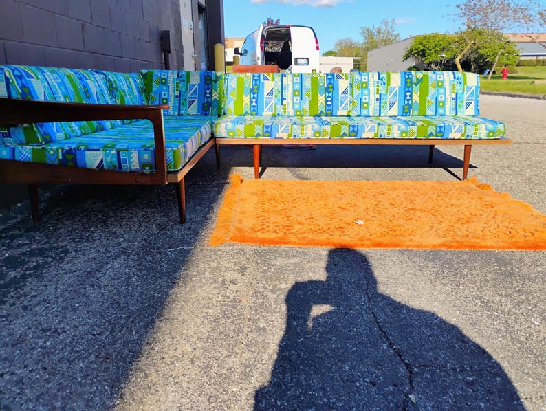 May include: A mid-century modern sectional sofa with a wooden frame and patterned cushions in shades of blue, green, and white. An orange shag rug is in front of the sofa. The sofa is outdoors on a sunny day.