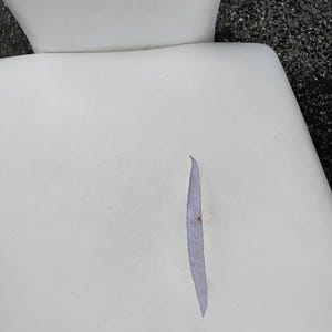 May include: Close-up of a white chair seat with a long, narrow tear. The tear reveals a lighter-colored interior. The chair's back is visible in the background, also white.
