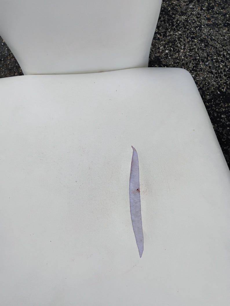 May include: Close-up of a white chair seat with a long, narrow tear. The tear reveals a lighter-colored interior. The chair's back is visible in the background, also white.