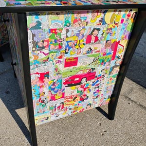 May include: A black-framed cabinet with a comic book collage design. The cabinet's surface and sides are covered in colorful comic strips, featuring various characters and scenes. The legs are black.