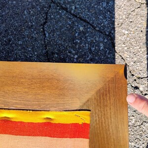 May include: A corner of a wooden framed artwork. The frame is a light brown color. The artwork has a colorful fabric design with yellow, orange, and red stripes. The artwork is on a cracked asphalt surface.