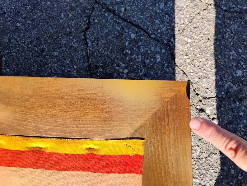 May include: A corner of a wooden framed artwork. The frame is a light brown color. The artwork has a colorful fabric design with yellow, orange, and red stripes. The artwork is on a cracked asphalt surface.