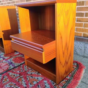 May include: Two wooden nightstands with a warm, reddish-brown finish. Each nightstand features a drawer and an open shelf. The nightstands are placed on a colorful rug with a geometric pattern. The wood grain is visible.
