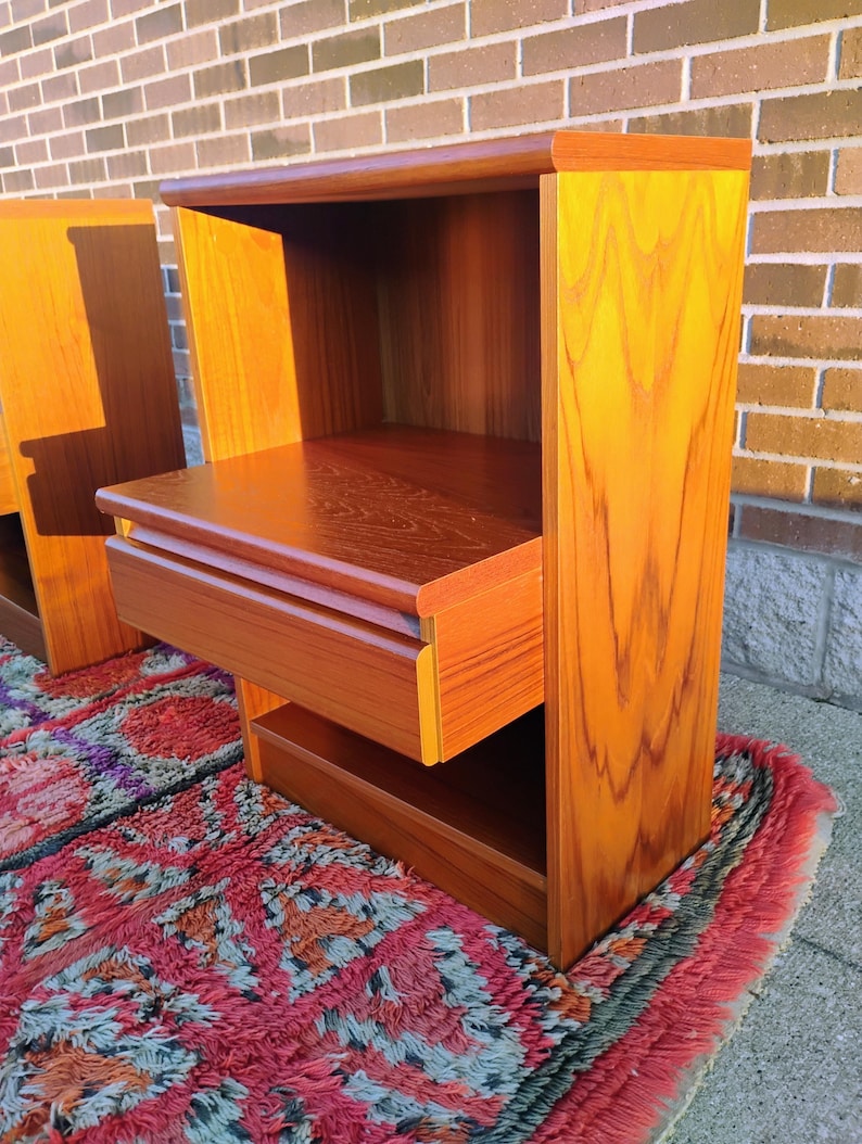 May include: Two wooden nightstands with a warm, reddish-brown finish. Each nightstand features a drawer and an open shelf. The nightstands are placed on a colorful rug with a geometric pattern. The wood grain is visible.