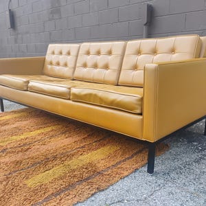 May include: A three-seat sofa upholstered in mustard yellow leather featuring a tufted backrest with buttons. The sofa has a rectangular frame, straight armrests, and is supported by a black metal base with four thin, vertical legs.