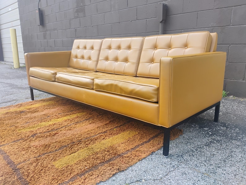 May include: A three-seat sofa upholstered in mustard yellow leather featuring a tufted backrest with buttons. The sofa has a rectangular frame, straight armrests, and is supported by a black metal base with four thin, vertical legs.