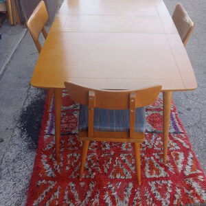 May include: A wooden dining table with four chairs. The table has a light wood finish and the chairs have a dark wood finish. The table and chairs are on a red and white rug.