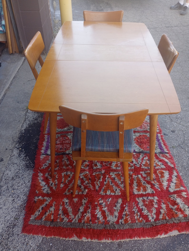 May include: A wooden dining table with four chairs. The table has a light wood finish and the chairs have a dark wood finish. The table and chairs are on a red and white rug.