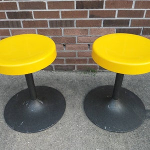May include: Two yellow round stools with black bases. The stools are made of plastic and metal.