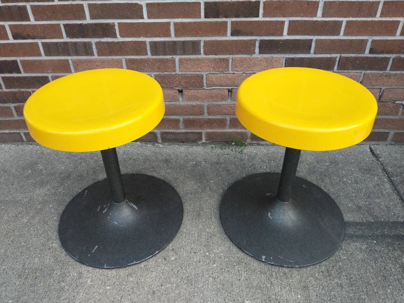 May include: Two yellow round stools with black bases. The stools are made of plastic and metal.