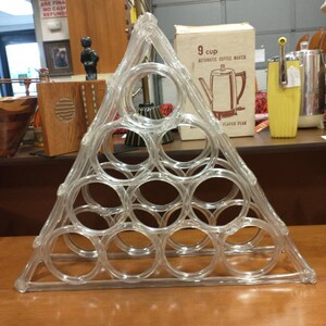 May include: A clear plastic triangular wine rack with 12 circular openings for holding wine bottles. The rack is on a wooden surface.